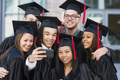 Multiracial group of six friends taking a selfie at graduation, in their black caps and gowns.